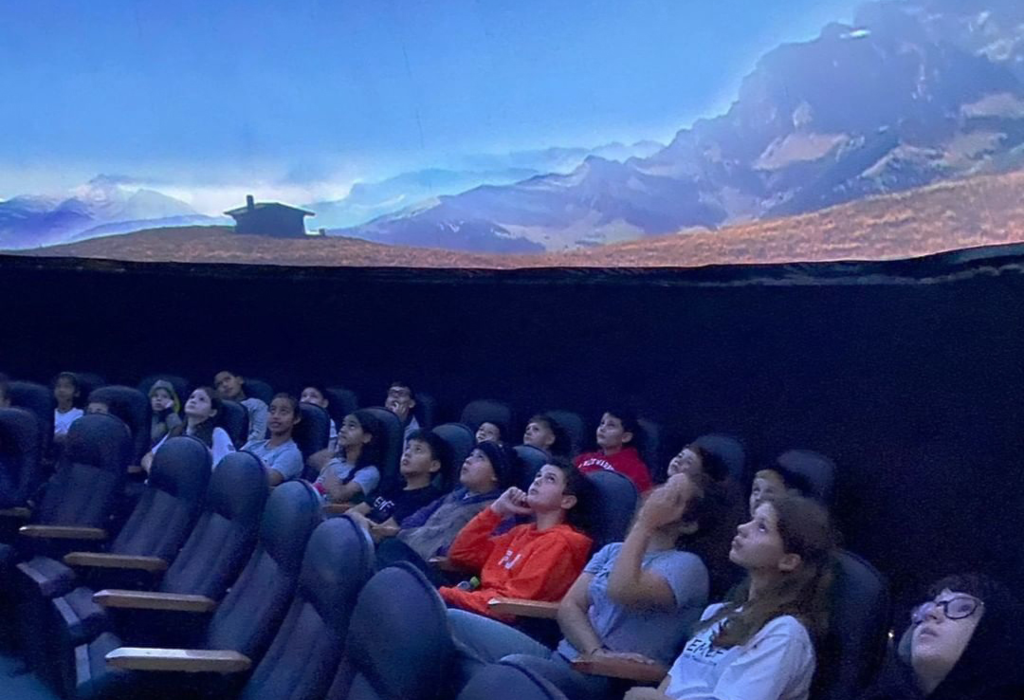 Crianças assistem a uma sessão no planetário, com projeção de céu azul no domo