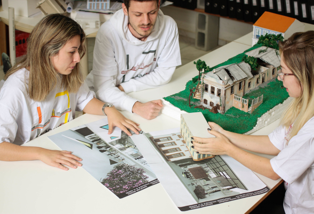 Três estudantes de Arquitetura e Urbanismo analisam fotos de prédios históricos ao lado de maquete