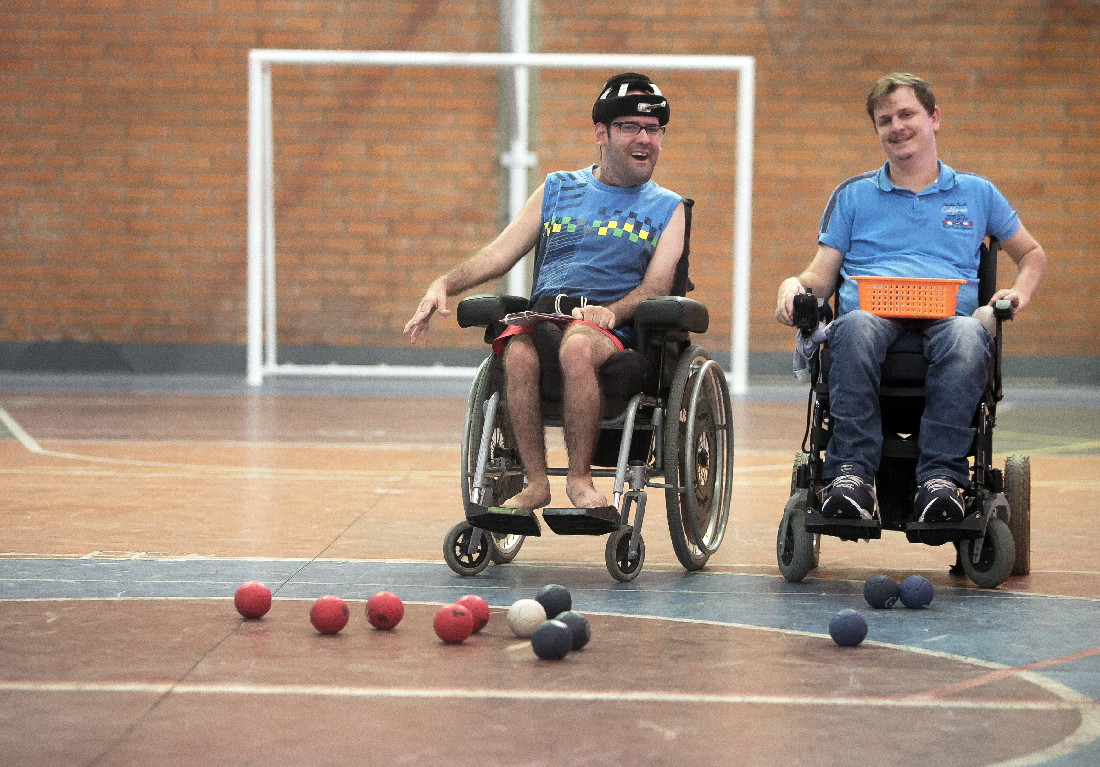 Ambiente interno, ginásio, dois meninos em cadeiras de rodas, algumas bolas no chão.