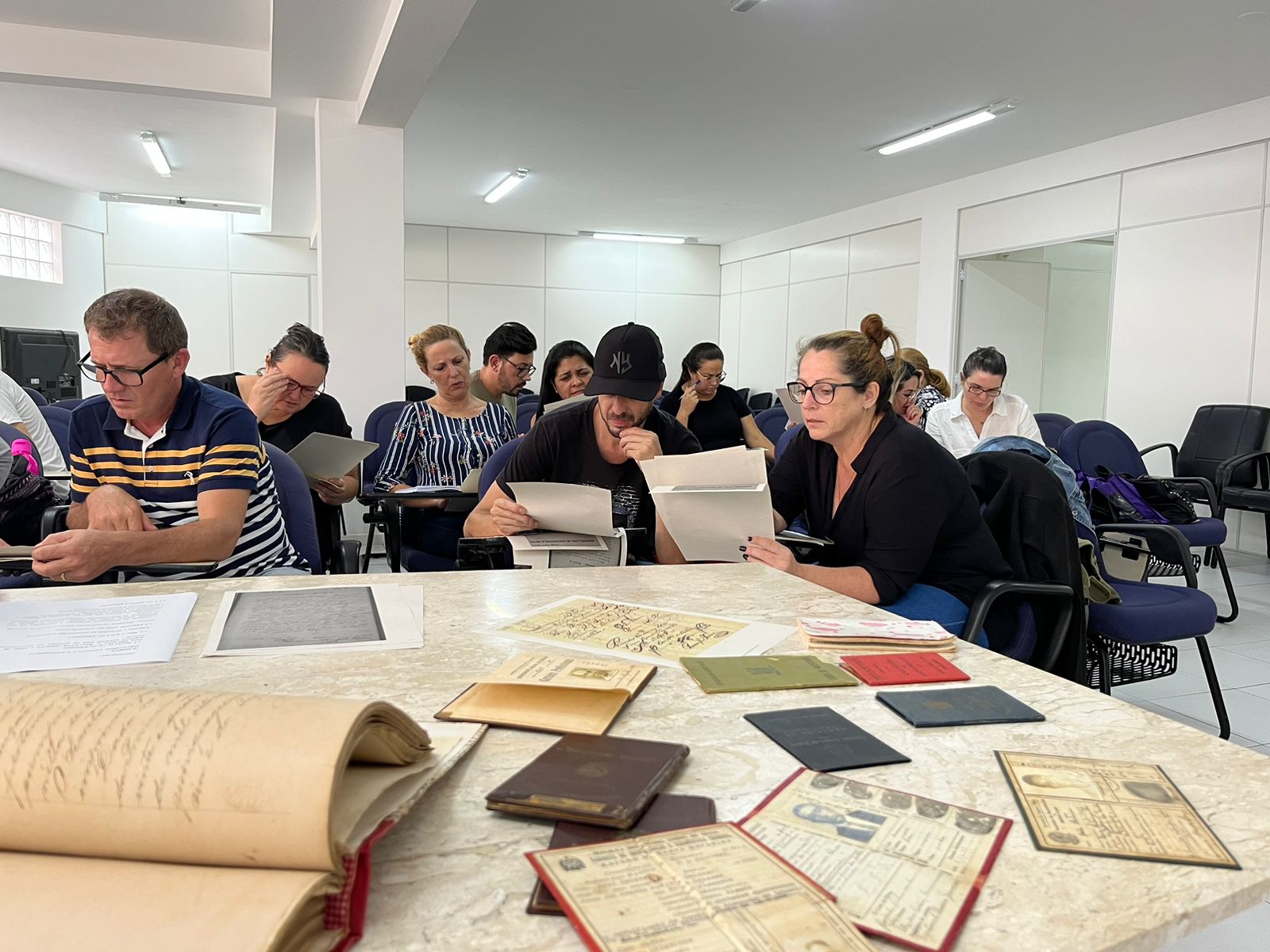 Livros e documentos históricos espalhados sobre mesa, enquanto professores, sentados em cadeiras, analisam registros históricos