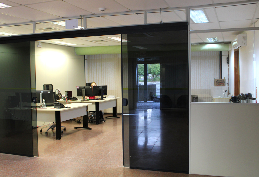 A foto mostra a entrada do Gabinete do Reitor, com portas de vidro duplas, mesas e computadores