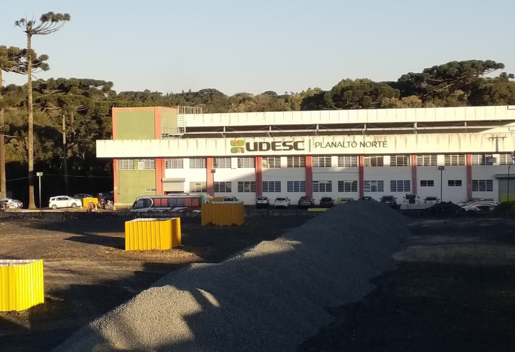 Imagem da área externa do campus com montes de brita e materiais de obra. Ao fundo, aparece o prédio da unidade.