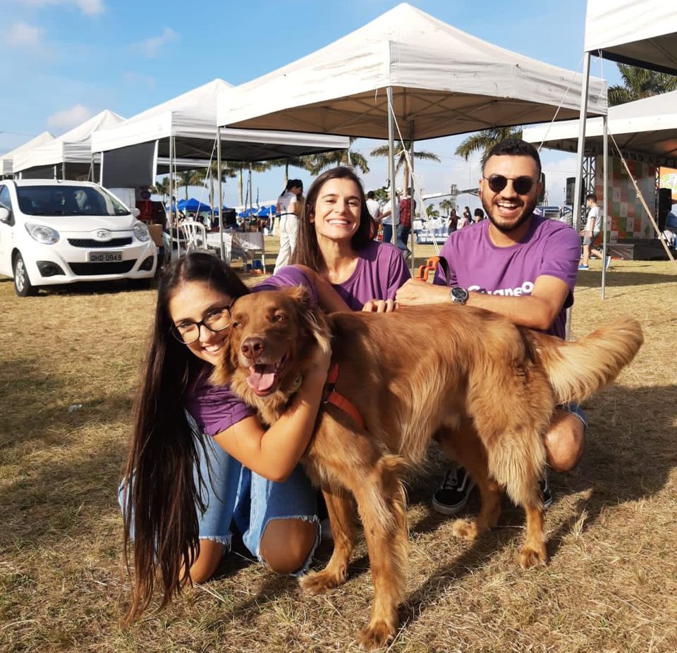 Fundadores da Guapeco, Julia Locks, Martha Rodrigues e Thiago Pimentel abraçam cão labrador em espaço gramado na Avenida Beira Mar Norte, em Florianópolis, com tendas de evento ao fundo.