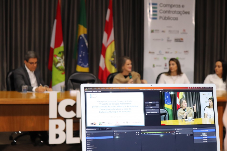 Participantes falam durante evento no Salão Nobre da Prefeitura de Bluemanau, com tela em primeiro plano do computador para controle da transmissão pelo Youtube
