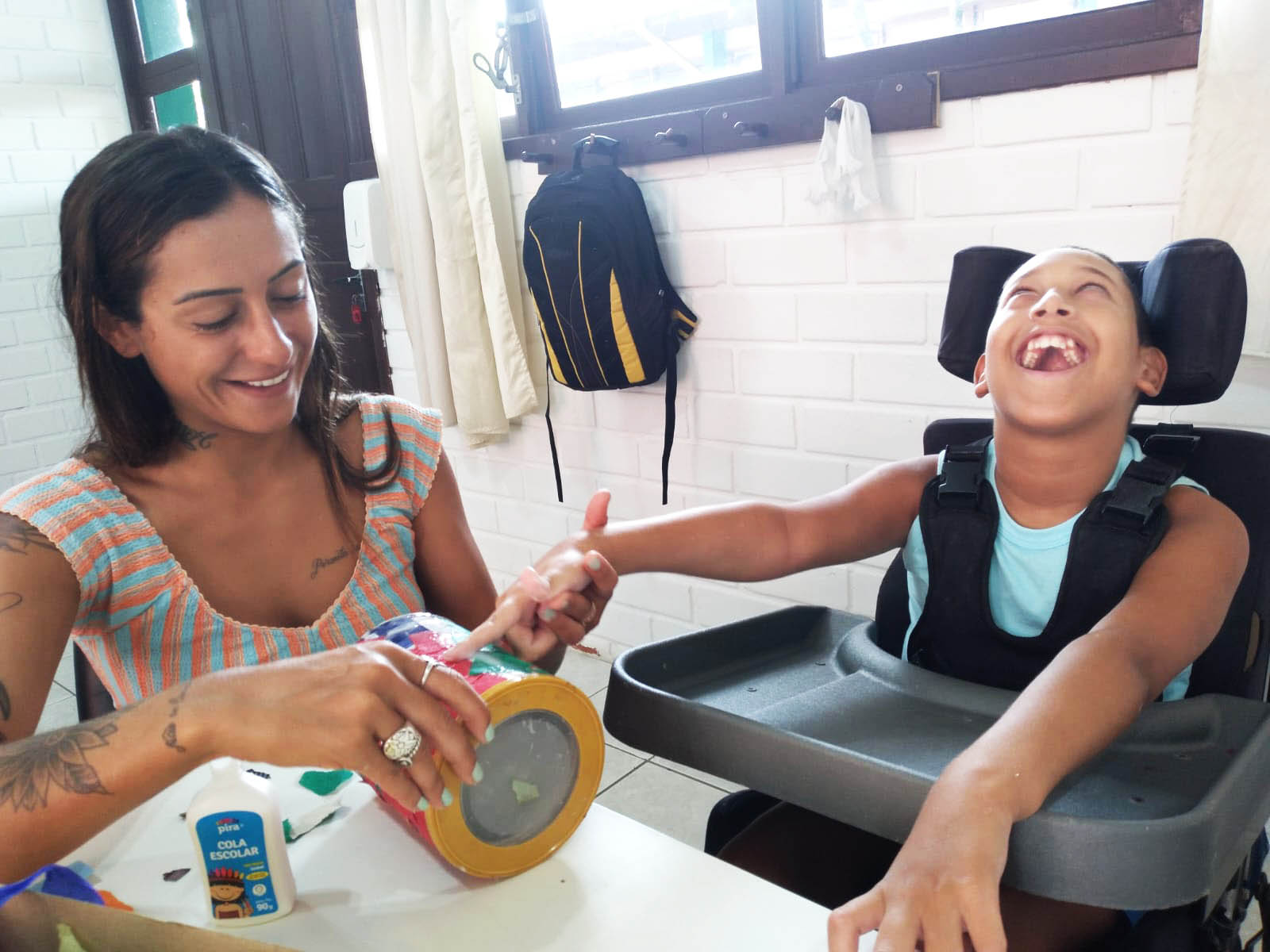 Duas pessoas sorrindo sentadas, uma mulher mexe com a mão de um menino em cadeira de rodas. ambiente interno.