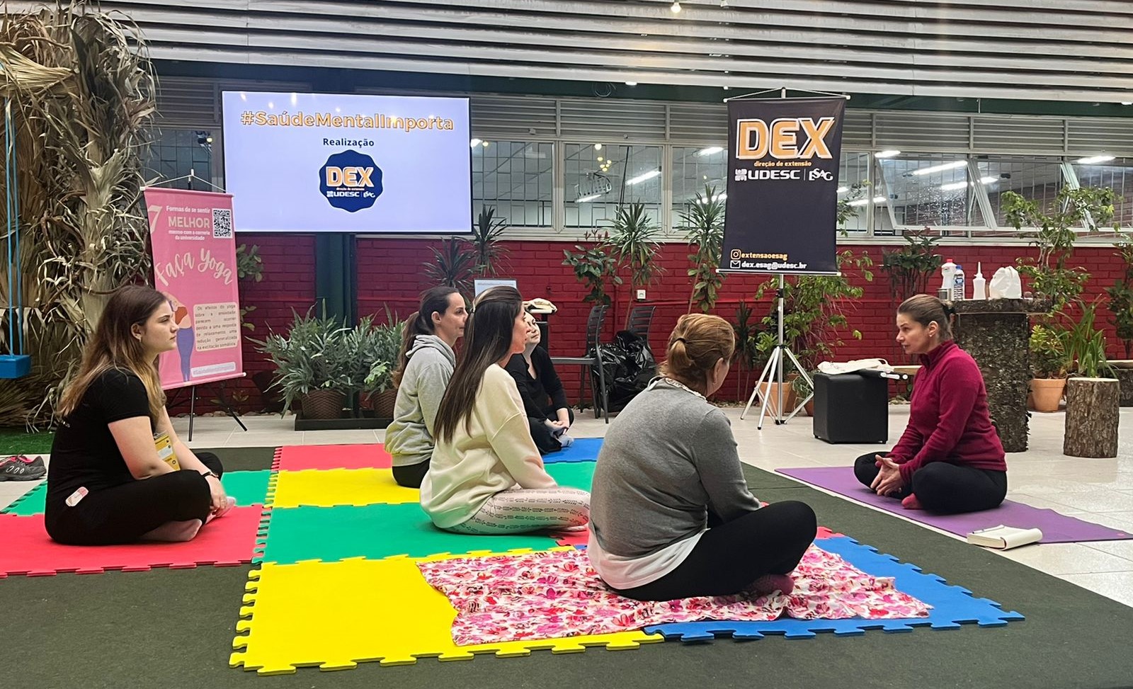 Grupo sentado no chão, sobre um tatame, em espaço decorado com plantas, recebendo instruções de professora de ioga, sentada de frente para o grupo de mulheres