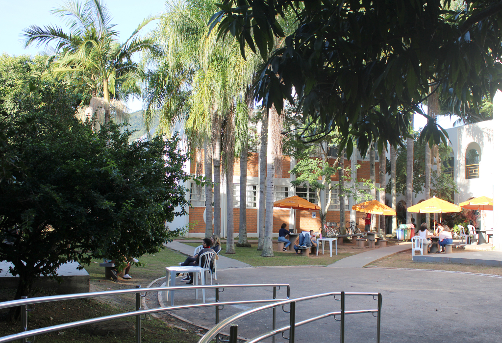 Foto do campus da Udesc em Florianópolis, com árvores e estudantes em mesas ao ar livre.