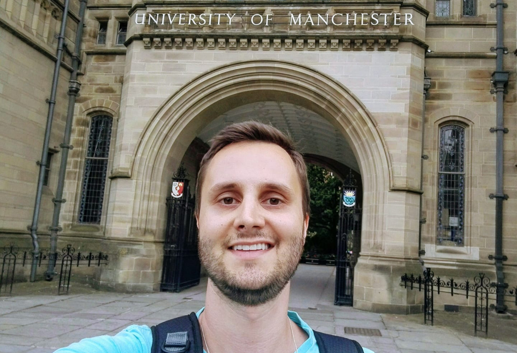 Foto do professor Marcelo, um homem branco, de cerca de 35 anos, com cabelo claro e barba. Atrás aparece a entrada da Universidade de Manchester, no Reino Unido.