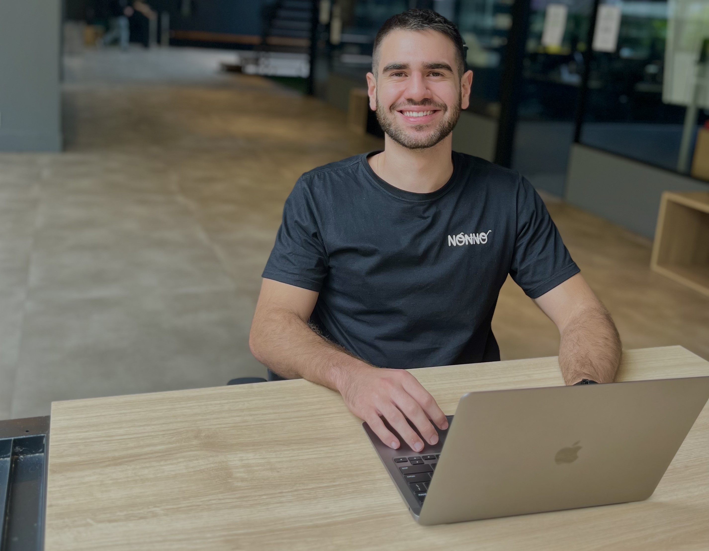 CEO da startup Nonno, Matheus Alban, de camiseta escura com a marca da empresa pequena estampada no lado esquerdo do peito, sentado em frente a uma mesa e trabalho com seu notebook, em ambiente corporativo.