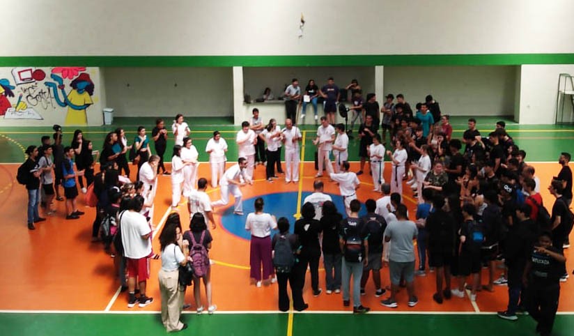 imagem de ginasio com roda com cerca de 80 pessoas, ao centro praticantes de capoeira vestidos de branco