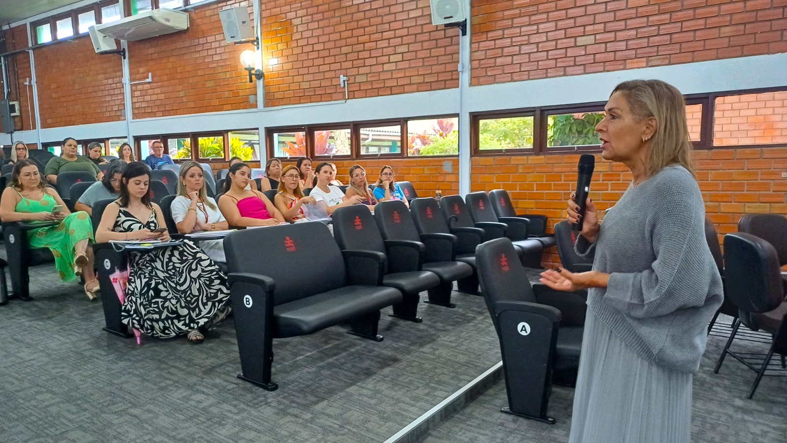 mulher em pé fala ao microfone, atrás pessoas sentadas em plateia de auditorio