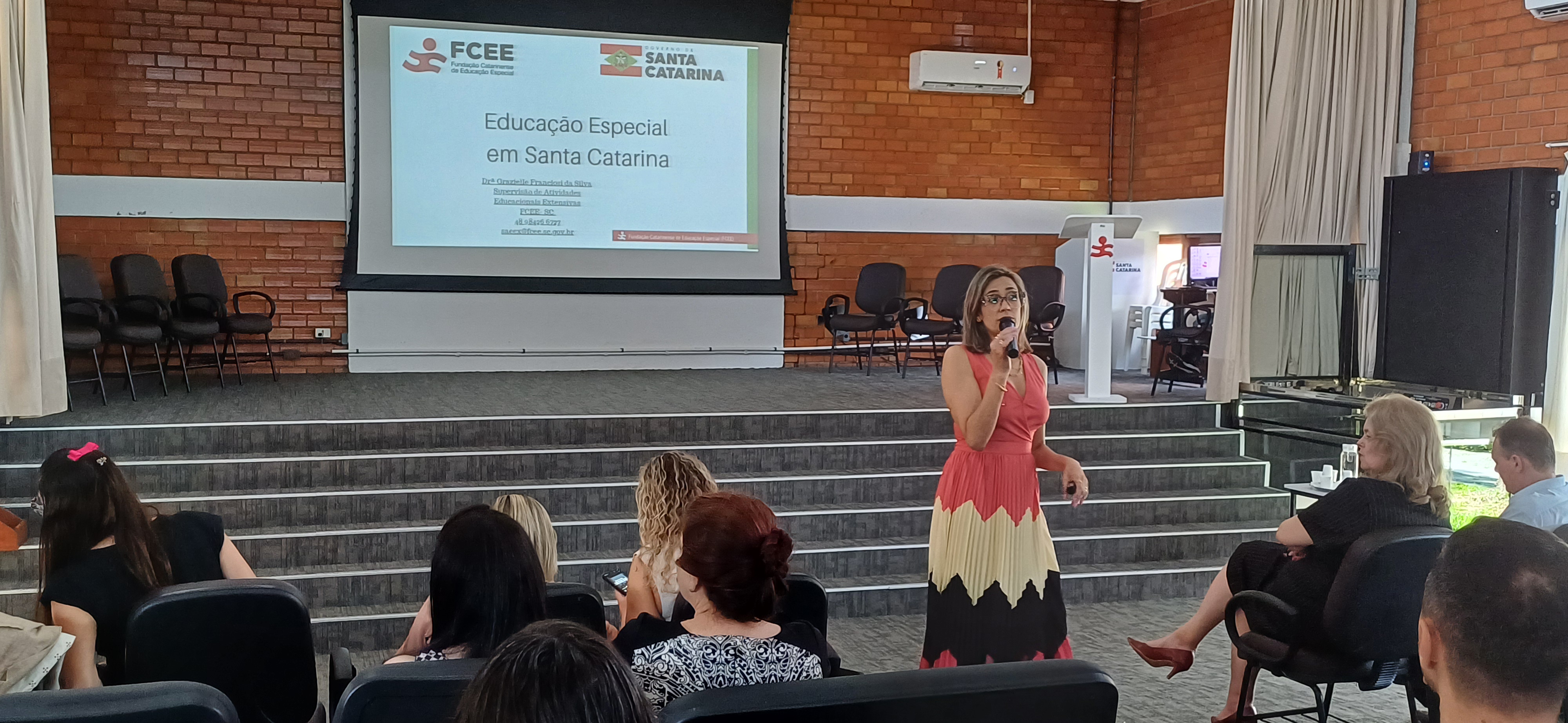 Mulher em pé com microfone fala diante de plateia em auditorio.